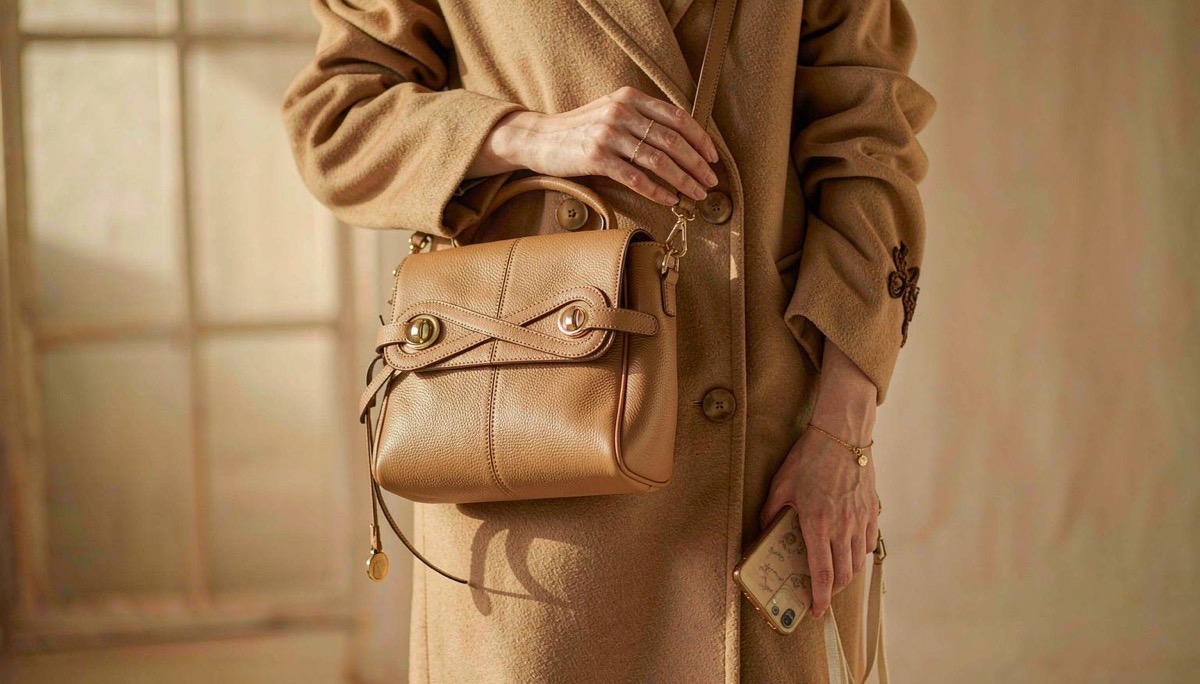 A tan leather shoulder bag with a long strap riding at the hip over a camel wool coat